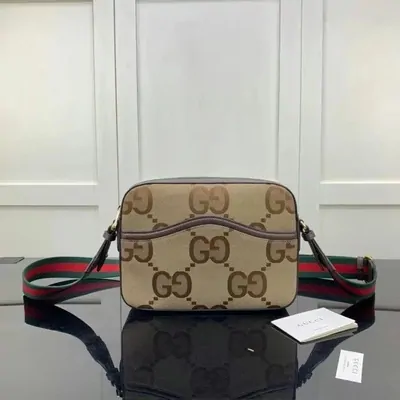 A monogram canvas crossbody bag with a red and green woven strap, photographed against a plain wall
