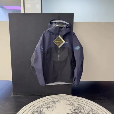A hooded waterproof shell jacket in grey and navy, hanging on a rail against a dark board