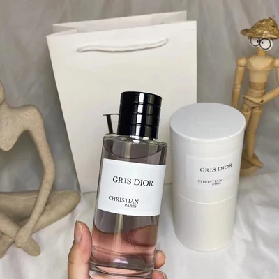 A hand holding a clear rectangular fragrance bottle of pale pink liquid with a plain white label, beside a white cylindrical box and a paper bag