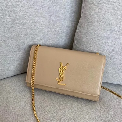 A beige grained leather flap wallet with a gold chain strap and a gold lettered plaque, resting against a cushion
